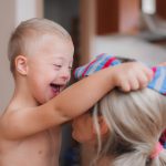 A happy child with Down syndrome, shirtless, laughing while playing with a striped fabric that they are placing on the head of a woman with blonde hair tied in a ponytail.