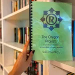Person holding a spiral-bound booklet titled 'The Oregon Project for Preschool Children Who are Blind or Visually Impaired: Skills Inventory,' with bookshelves in the background.
