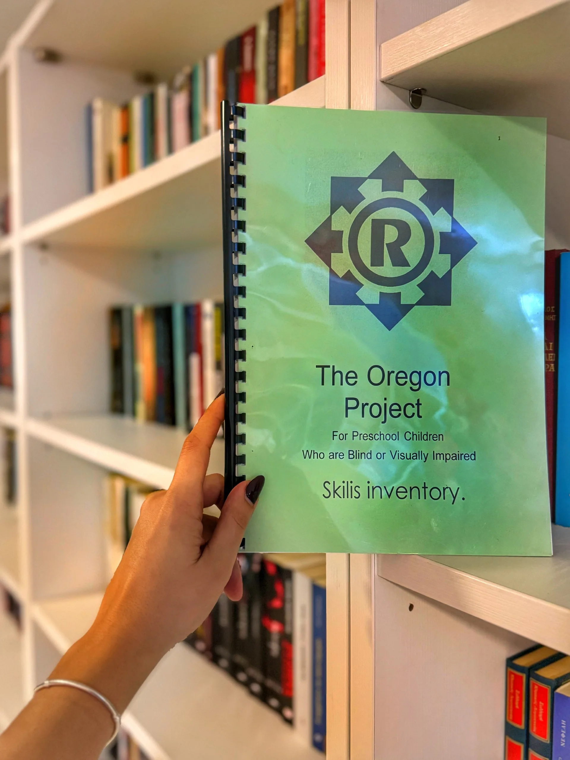 Person holding a spiral-bound booklet titled 'The Oregon Project for Preschool Children Who are Blind or Visually Impaired: Skills Inventory,' with bookshelves in the background.
